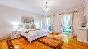 A bedroom with a large bed, a chandelier, a nightstand, and a view of greenery through the window.
