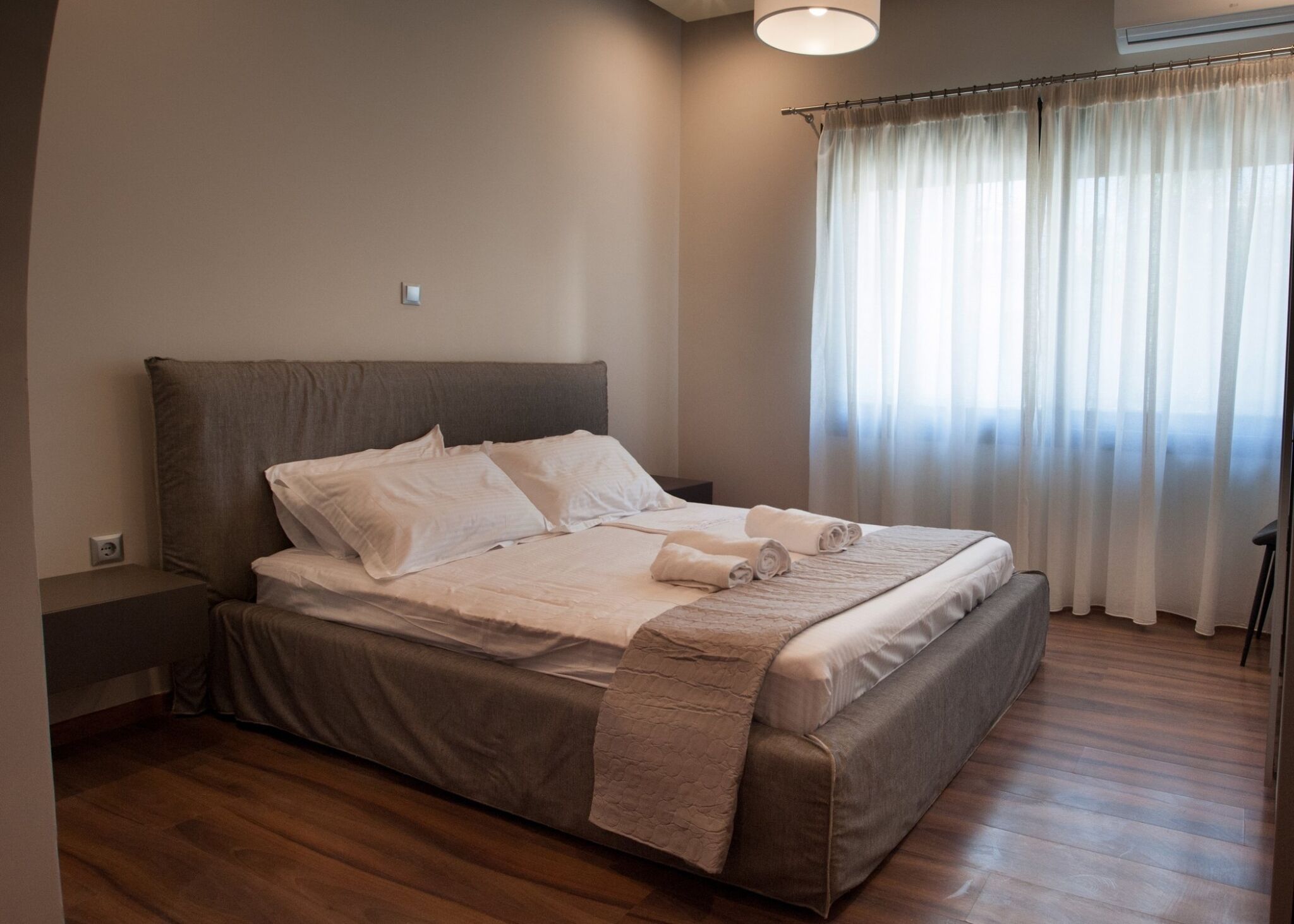 A neatly made bed with a brown headboard, a bedside table, and a window with sheer curtains.
