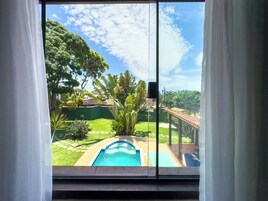 Vista pela janela com cortinas brancas, mostrando piscina, gramado verde e árvores ao redor.