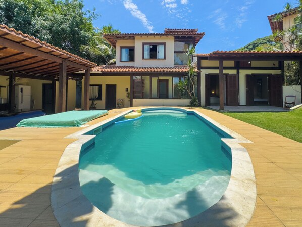 Piscina com deck ladrilhado, área de varanda coberta e um edifício de dois andares com telhado e janelas azulejadas.