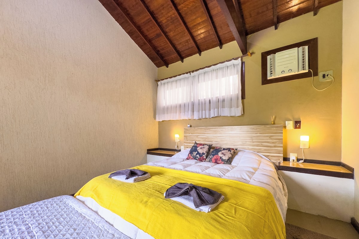 A hotel room with a bed, wooden headboard, bedside tables with lamps, a window with curtains, and a wall-mounted thermostat.