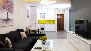 A modern living room with a black sofa, a coffee table with a vase and water bottles, a flat-screen TV, and a kitchen in the background with white cabinets and a yellow backsplash. - Welcome Northern Avenue Apartments (Yerevan)