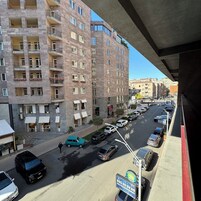 Een meerlaags woongebouw met balkons, een straat met geparkeerde auto's en een voetgangersoversteekplaats.