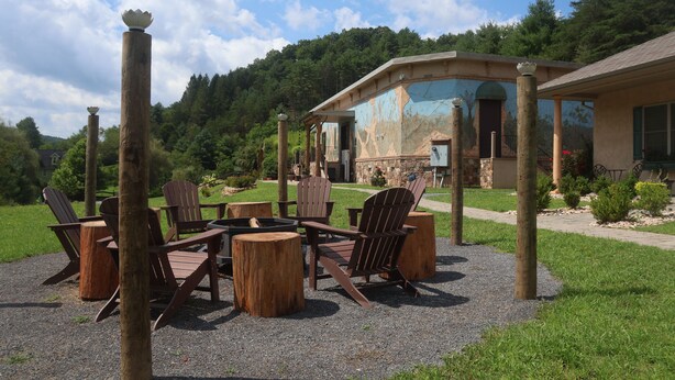 An outdoor seating area with Adirondack chairs and a fire pit, surrounded by wooden stumps, in front of a rustic building with a stone facade.