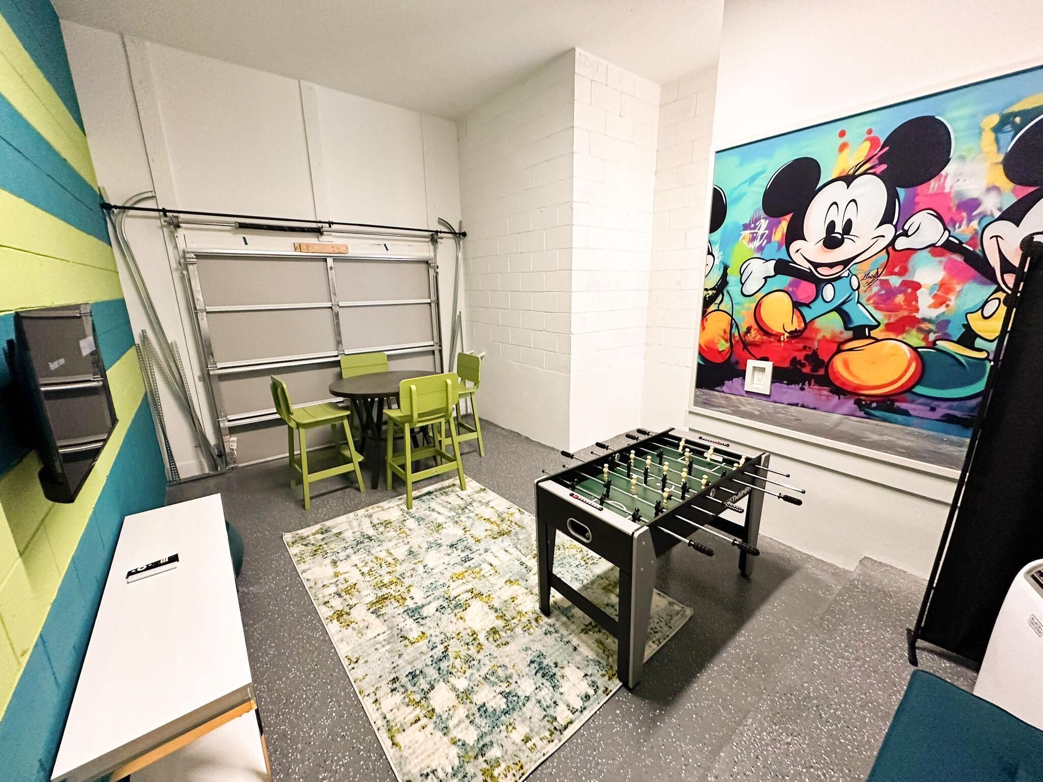 Una habitación con una mesa de futbolín, sillas verdes, una mesita y un mural colorido en la pared.