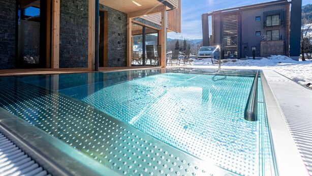 Una piscina tancada amb vidre i un pont de fusta, tot envoltat per una paret de pedra i un edifici de fusta.