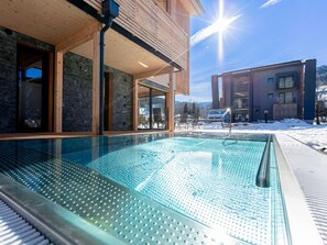 A glass-enclosed swimming pool with a wooden deck, surrounded by a stone wall and a wooden building. - Cosy Apartment With Sauna (Sankt Georgen am Kreischberg)