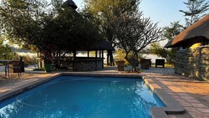 A swimming pool with a thatched-roof structure nearby, surrounded by trees and a stone wall. - Big 5 Toro Lodge (Kasane)