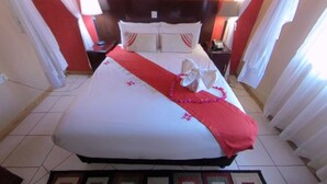 A large bed with a red bedspread and white pillows, a patterned rug, and a nightstand with a lamp. - Big 5 Toro Lodge (Kasane)