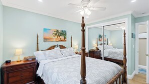 Une chambre à coucher avec un lit à baldaquin, un ventilateur de plafond, une table de chevet avec une lampe et un tableau représentant une scène de plage.