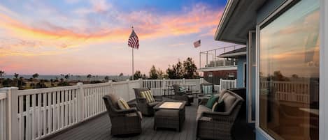 A deck with outdoor furniture, two American flags, and a view of the ocean at sunset.