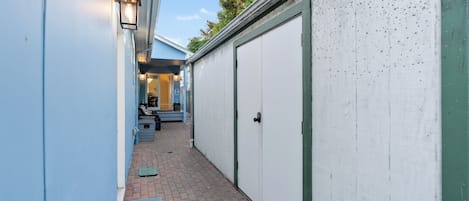 Ein schmaler Gang mit einem blauen Gebäude auf einer Seite und einem weißen Gebäude mit einer Garagentür auf der anderen. Es gibt Außenbereiche mit Sitzgelegenheiten wie Stühlen und Tischen, und eine kleine Stufe, die zu einer Tür führt.