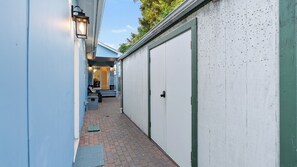 A narrow alleyway with a blue building on one side and a white building with a garage door on the other. There are outdoor seating areas with chairs and tables, and a small step leading to a door.