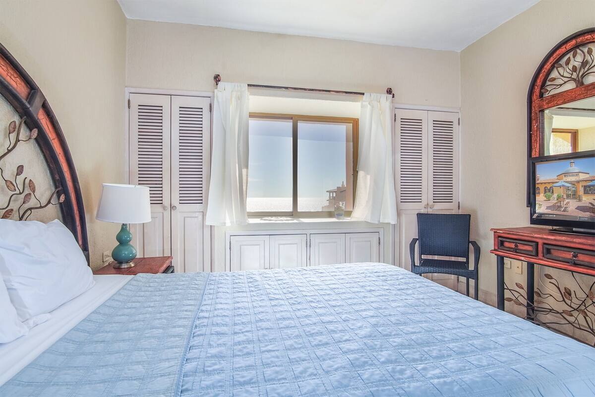 A hotel room with a bed, a chair, a desk with a television, and a window with shutters.