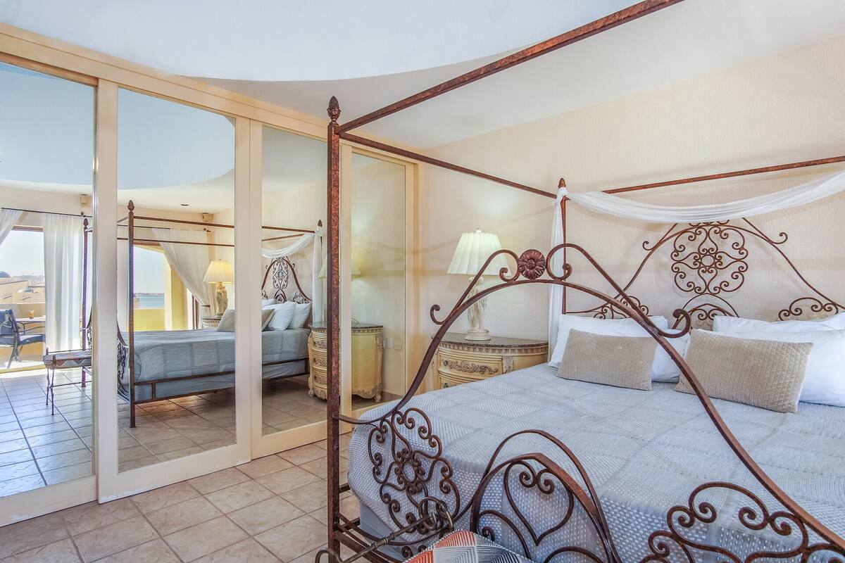 A four-poster bed with a canopy, a bedside table with a lamp, and a view through a glass door to an outdoor seating area.