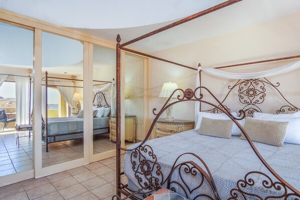 Una cama con dosel y cortinas, una mesita de noche con lámpara, y vista a través de un ventanal a un área de estar exterior.