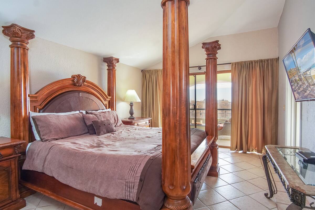 A four-poster bed with a wooden headboard, a bedside table with a lamp, and a view of the city through large windows.
