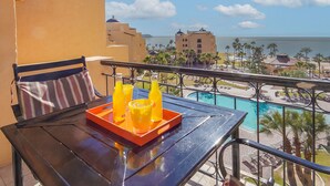Ein Balkon mit Tisch, zwei Stühlen, einem gestreiften Kissen und einer Ablage mit zwei Orangensaftgläsern und einer Flasche.