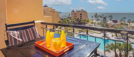 Een balkon met een tafel, twee stoelen, een gestreept kussen en een dienblad met twee glazen sinaasappelsap en een fles.