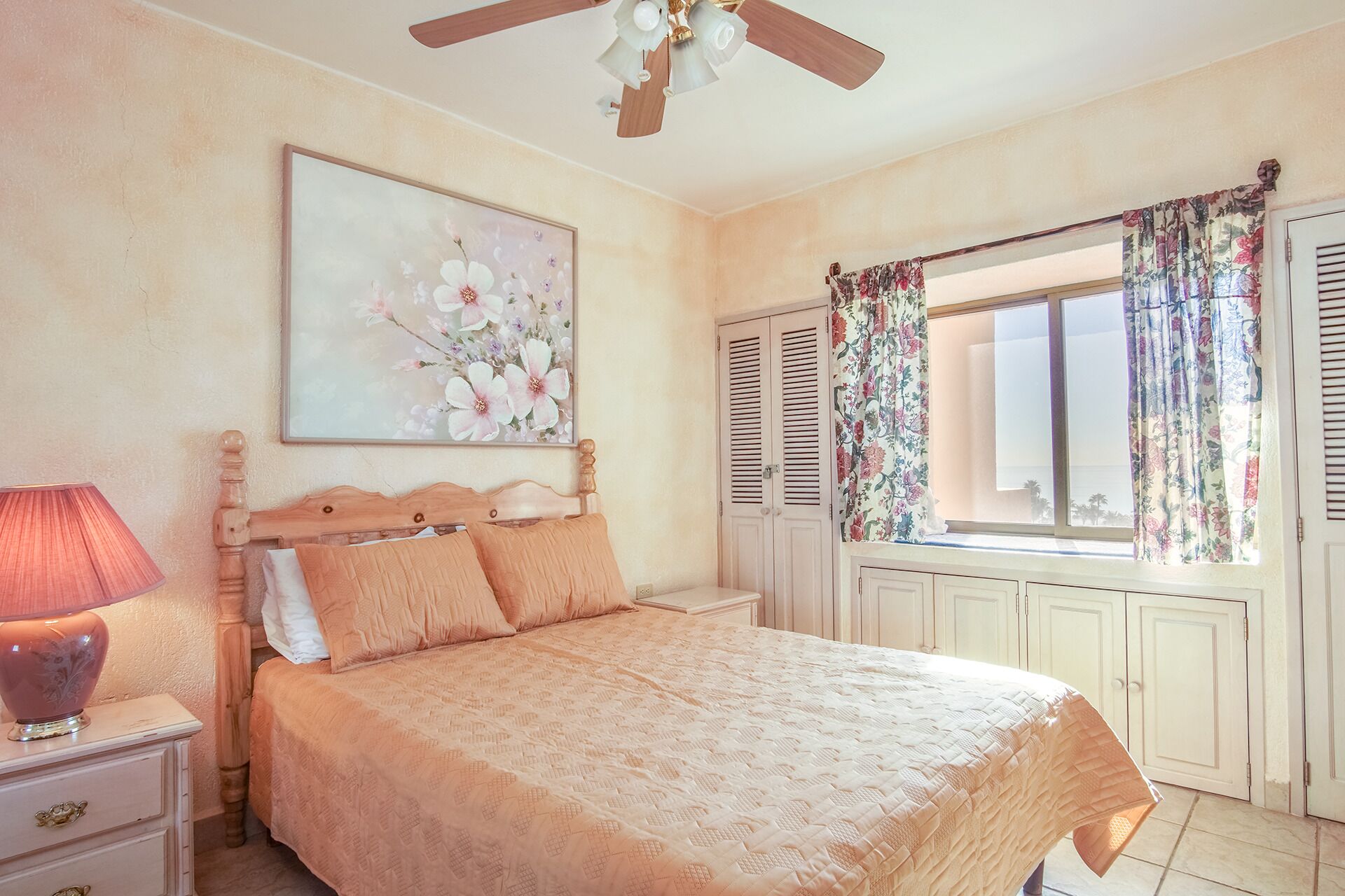 A bedroom with a bed, a nightstand, a lamp, a ceiling fan, a window with curtains, and a floral painting on the wall.