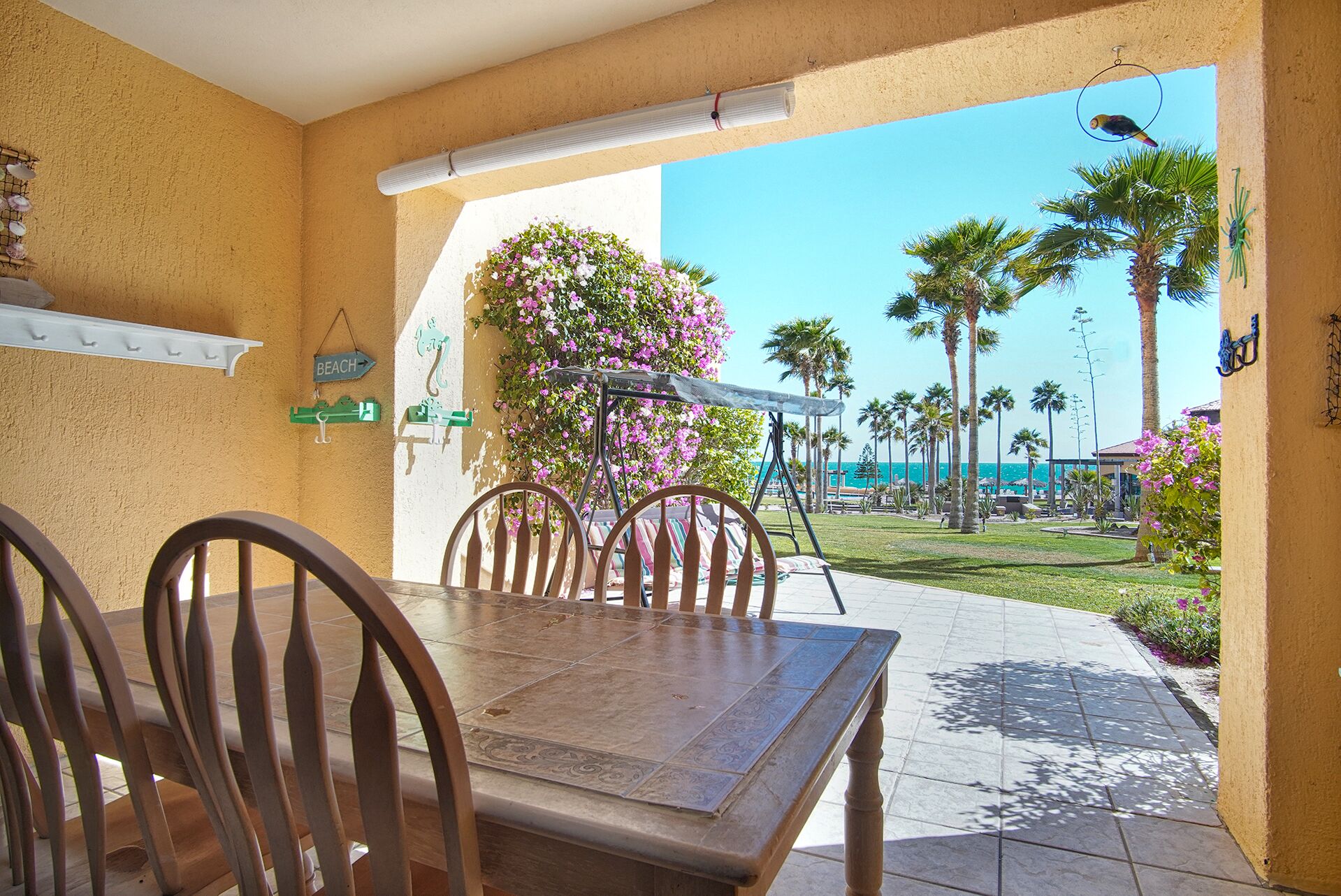 A patio with a wooden table and chairs, a hanging swing, and a view of palm trees and the ocean.
