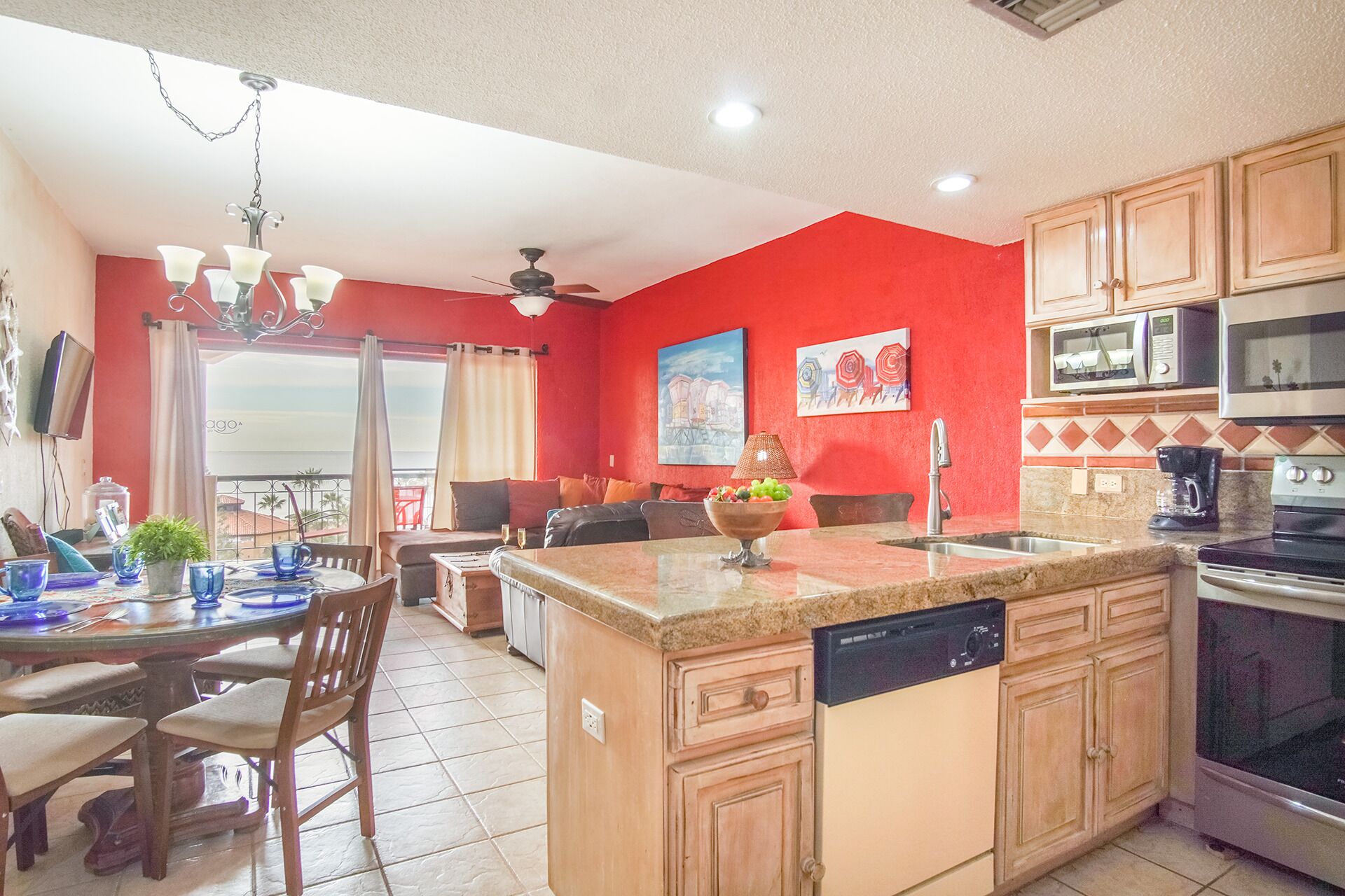 A kitchen with wooden cabinets, a microwave, an oven, and a dishwasher. There is a dining area with a table and chairs, and a balcony with a view.