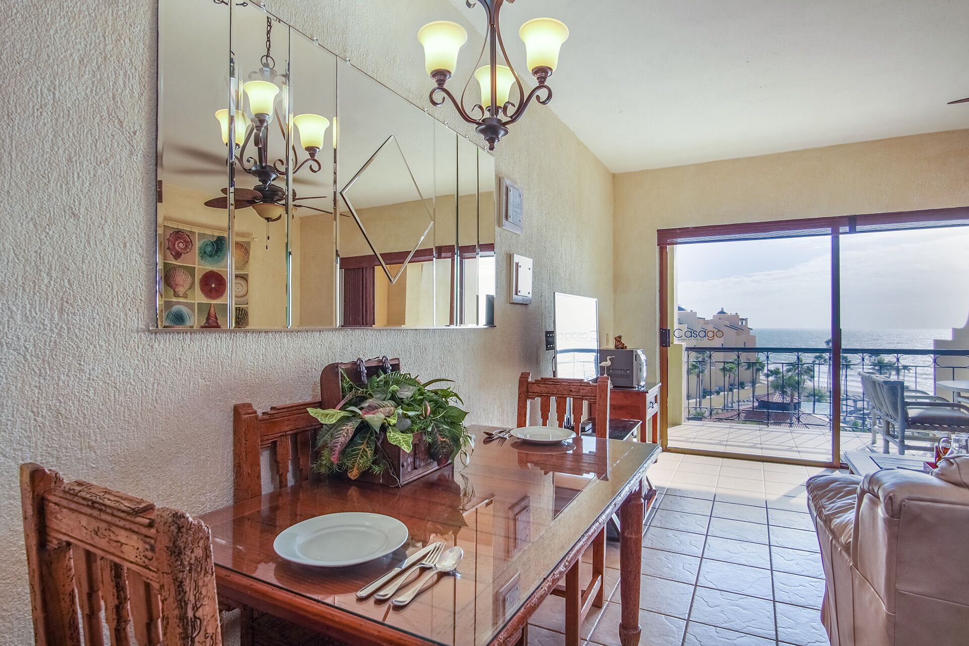 Un comedor con mesa de madera, sillas y un candelabro. Un espejo grande refleja la habitación, y se observa un balcón con vista al océano.