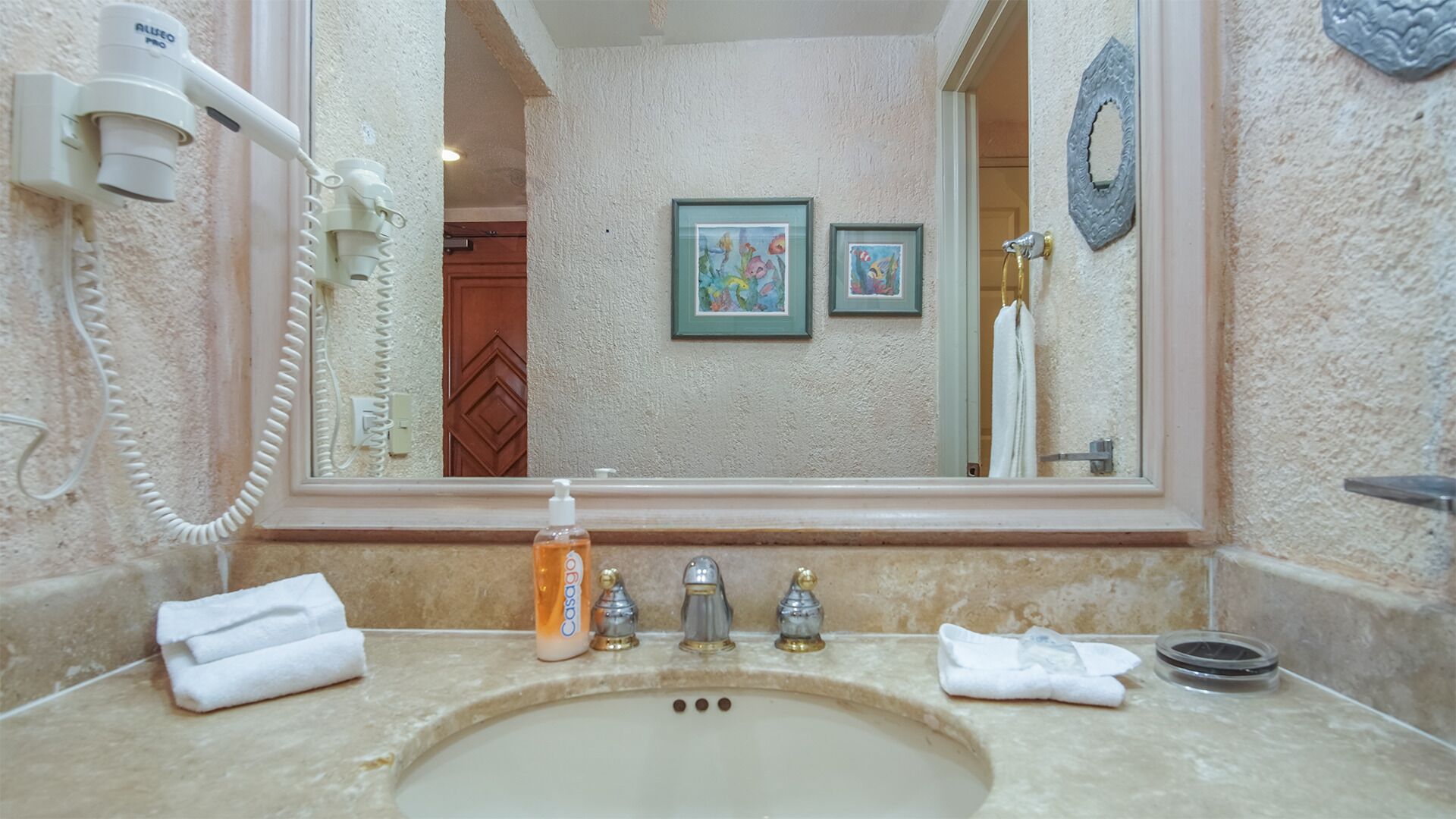 Un bagno con un lavandino, un asciugacapelli, una boccetta di sapone e due quadri appesi al muro.