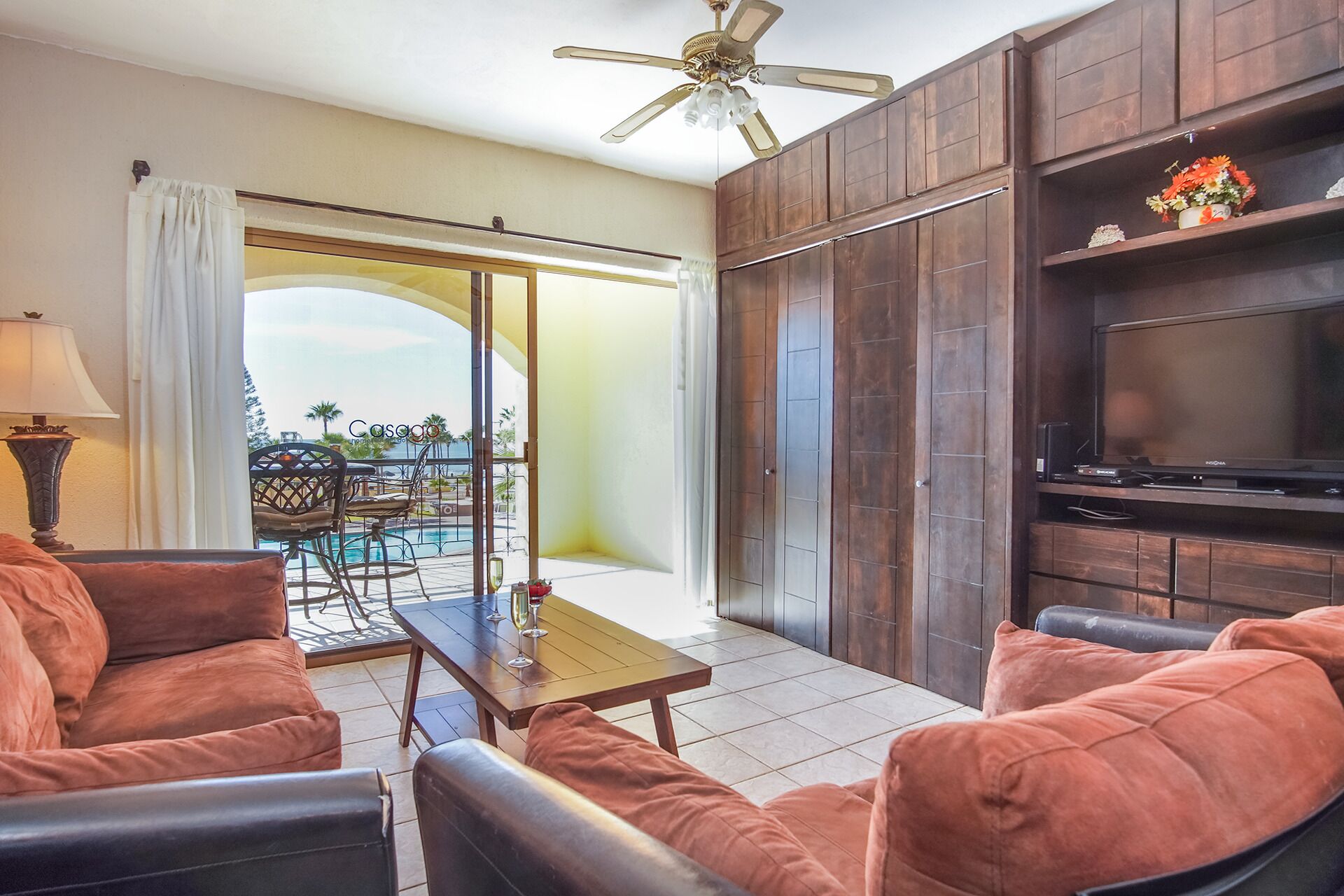 A living room with a TV, wooden cabinets, and a balcony with a pool view.