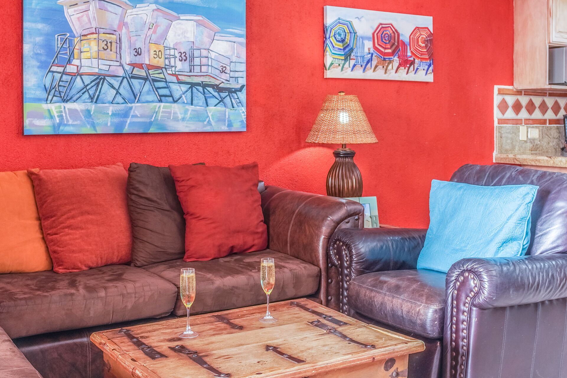 Ein Wohnzimmer mit einem braunen Ledersofa, einem Holztisch und einer roten Wand. Zwei Gemälde hängen an der Wand, und auf einem Beistelltisch steht eine Lampe.