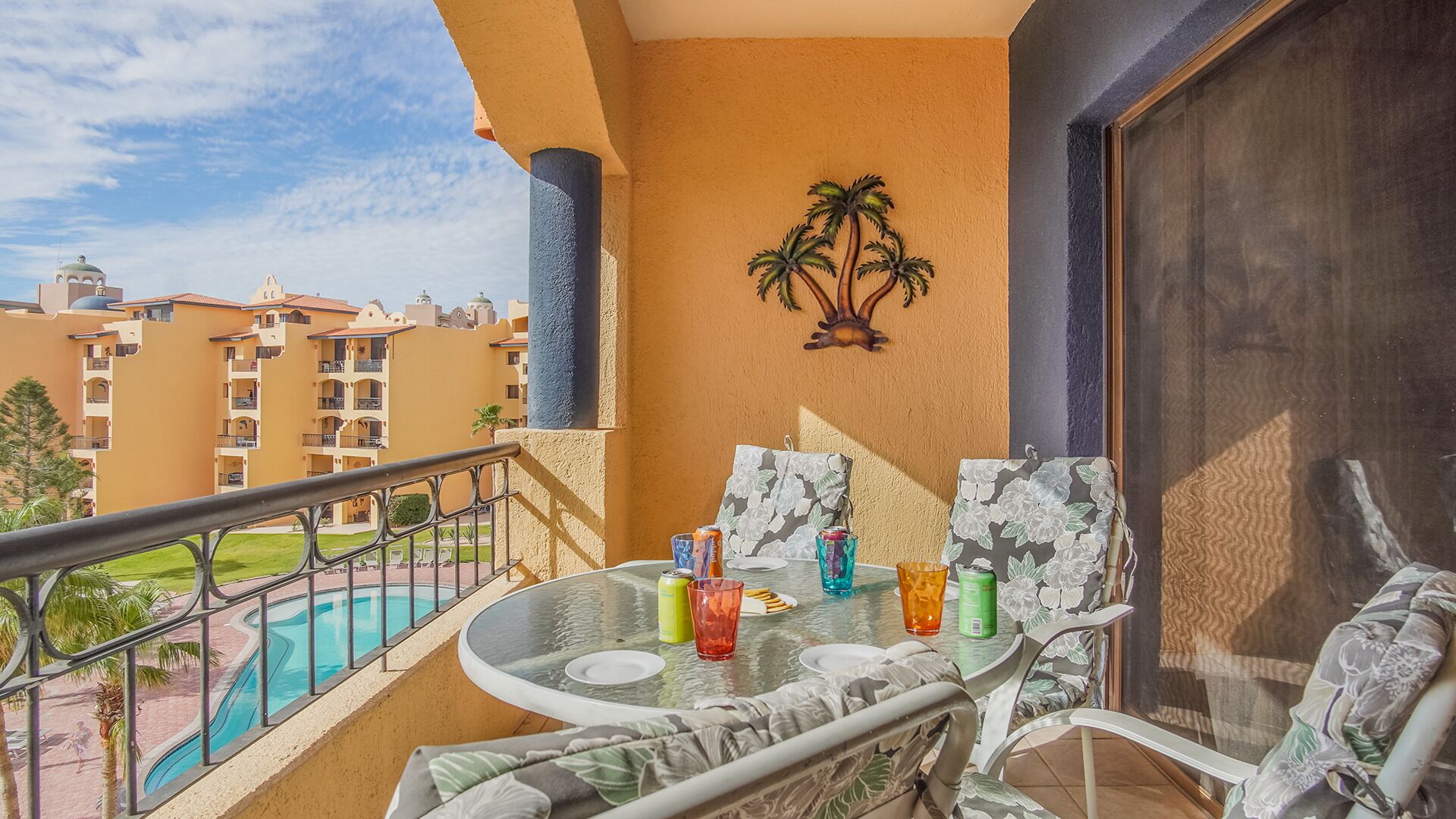 Un balcón con una mesa de vidrio, dos sillas, un cuadro de pared con motivos de palmera y vistas a una piscina y edificios de hotel.