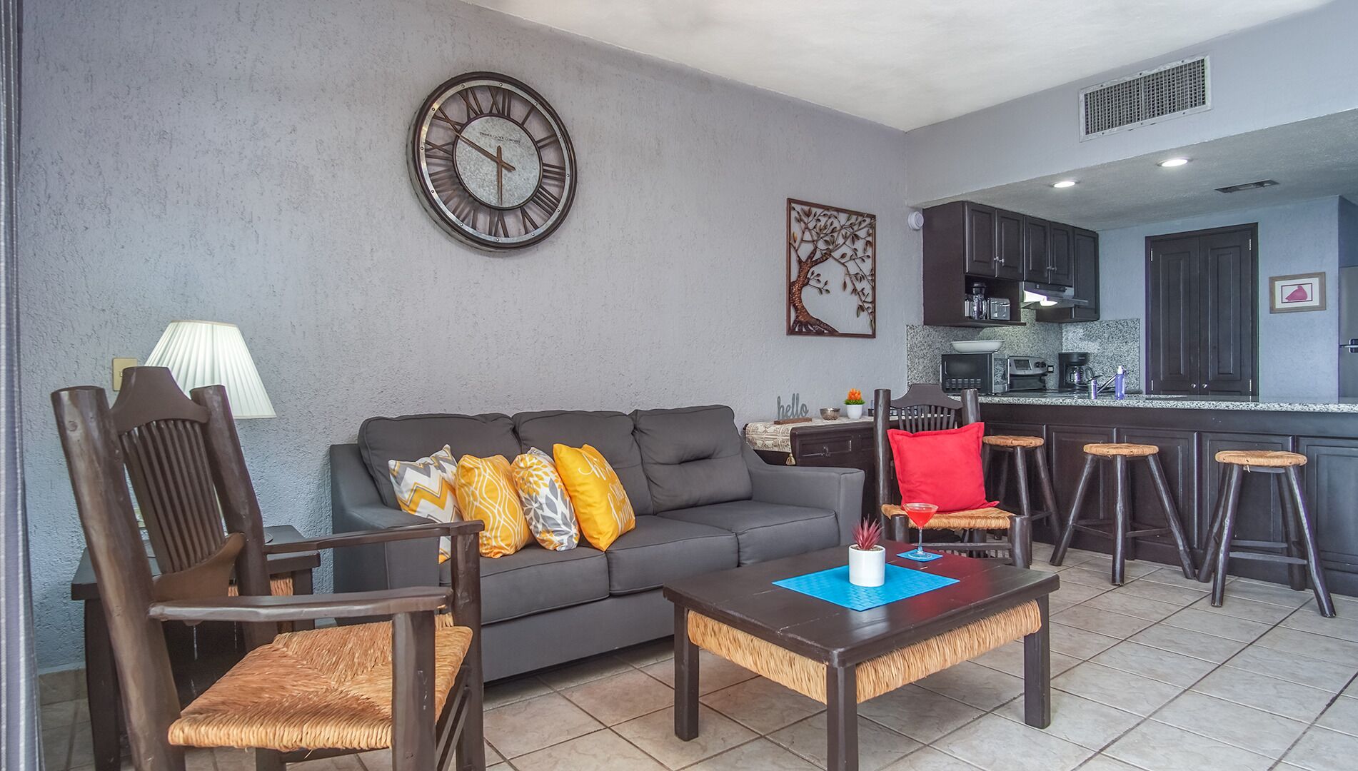 A living room with a grey sofa, wooden chairs, a coffee table, a clock, and a kitchen area with dark cabinets and a bar counter.