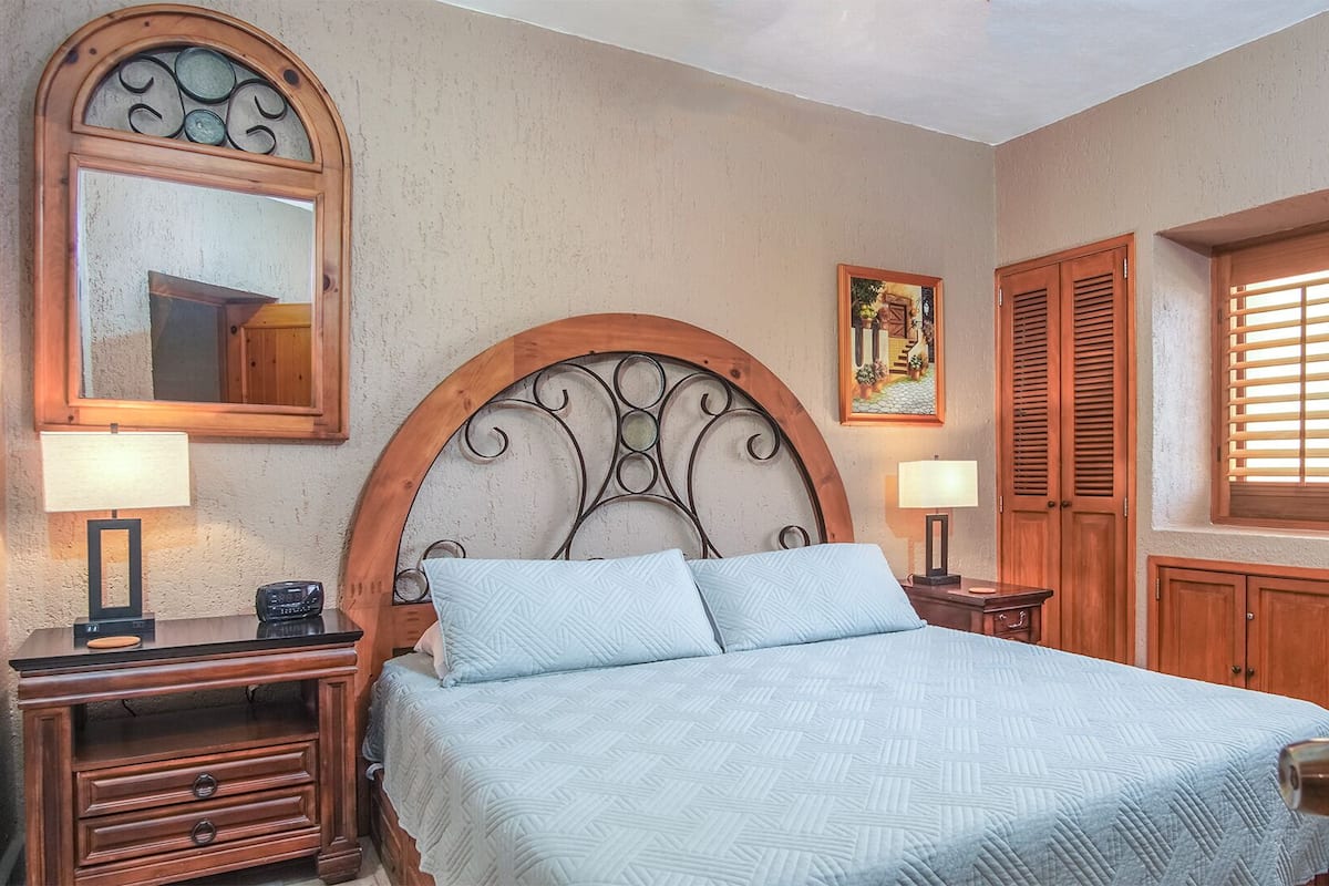 A bedroom with a large bed, two bedside tables, a wooden headboard, a mirror, a painting, and a window with shutters.