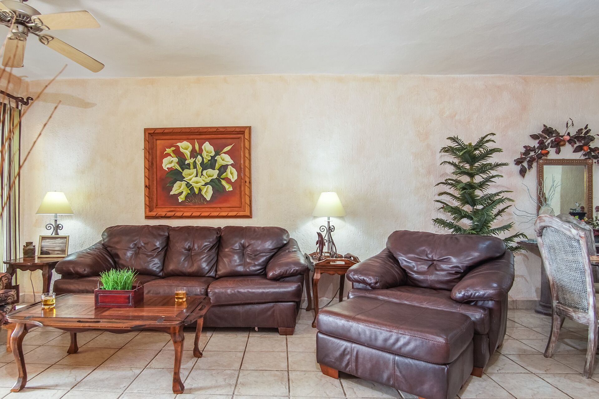 Una sala de estar con dos sofás de cuero, una mesa de centro de madera, un cuadro floral enmarco, una planta en maceta y una planta verde alta.