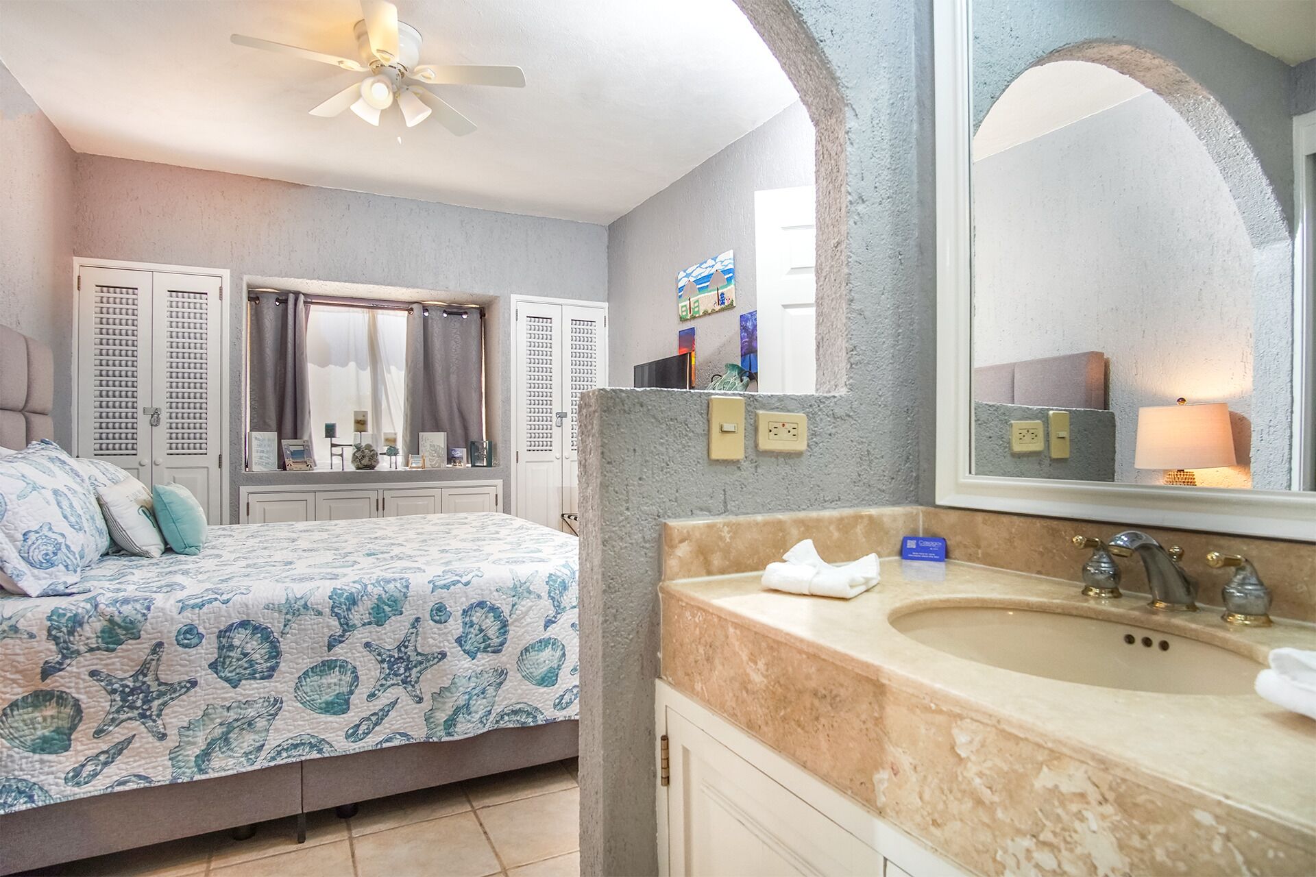 A bedroom with a bed, a ceiling fan, a window with curtains, and a bathroom with a sink and mirror.