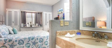 A bedroom with a bed, a ceiling fan, a window with curtains, and a bathroom with a sink and mirror.