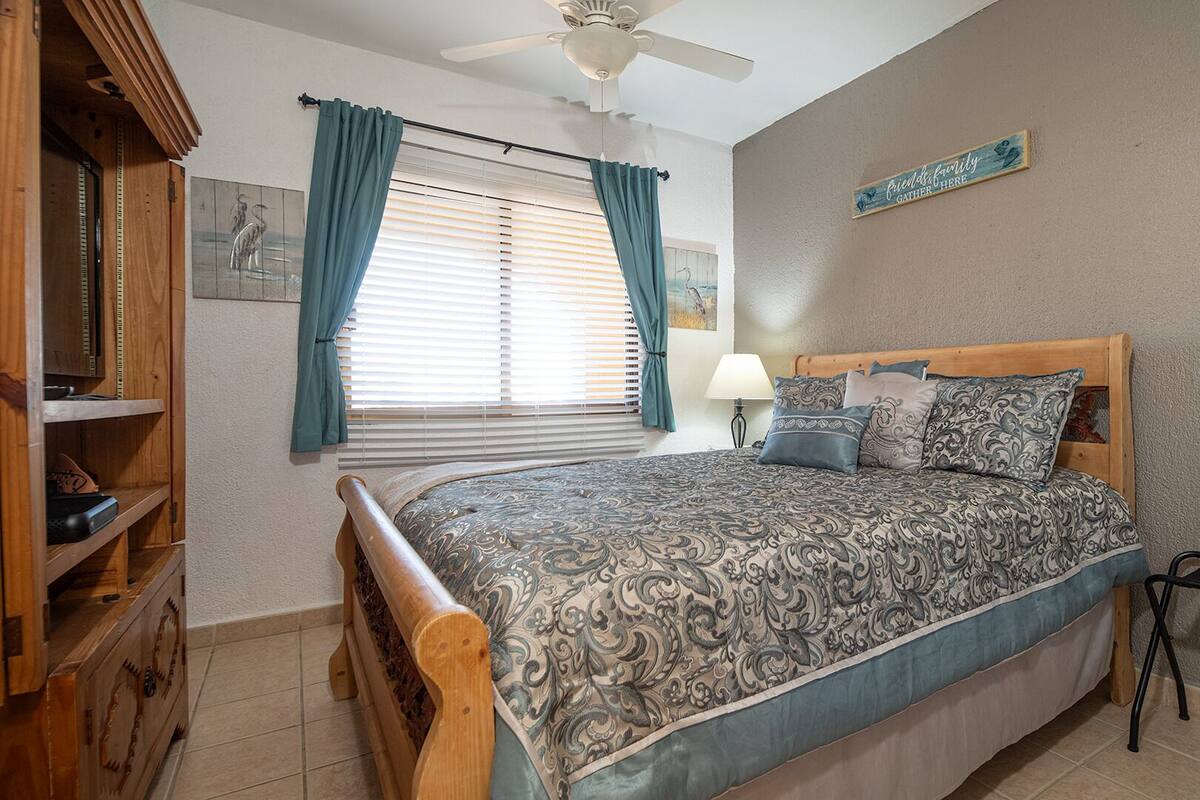 A bedroom with a bed, a wooden headboard, a ceiling fan, a window with curtains, a wall-mounted sign, and a small shelf with items.
