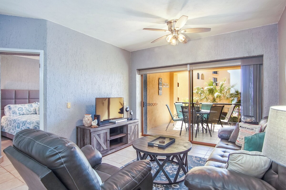A modern living room with a sofa, coffee table, and a TV. There is a balcony with a table and chairs.