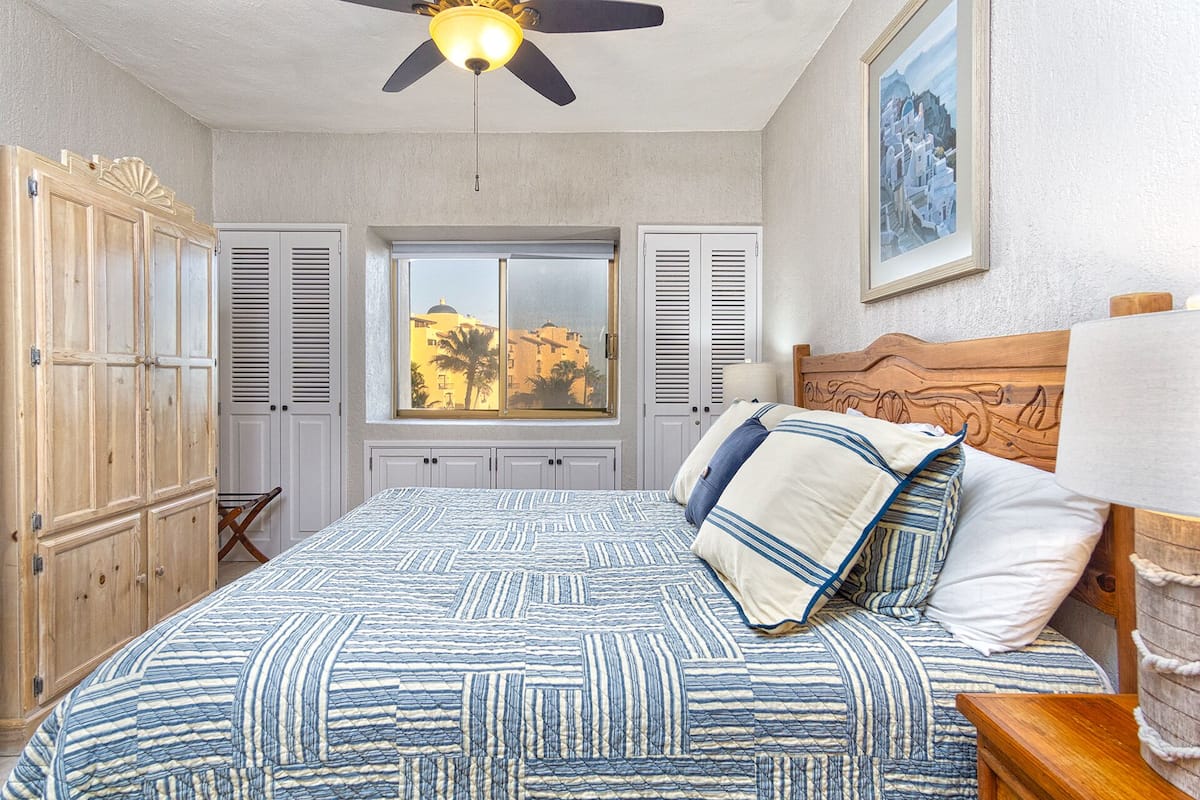 A bedroom with a bed, wooden headboard, a ceiling fan, a window with shutters, and a framed picture on the wall.