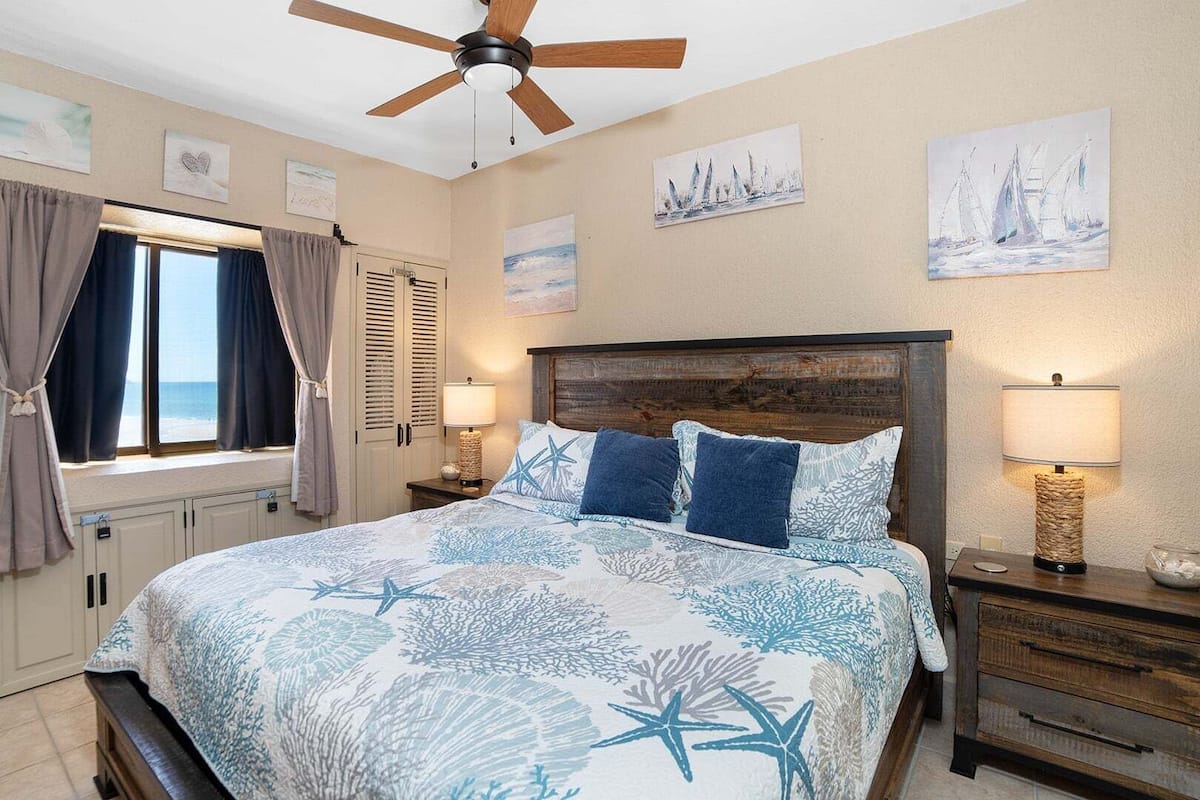 A bedroom with a bed, a ceiling fan, artwork on the walls, and a window with curtains.