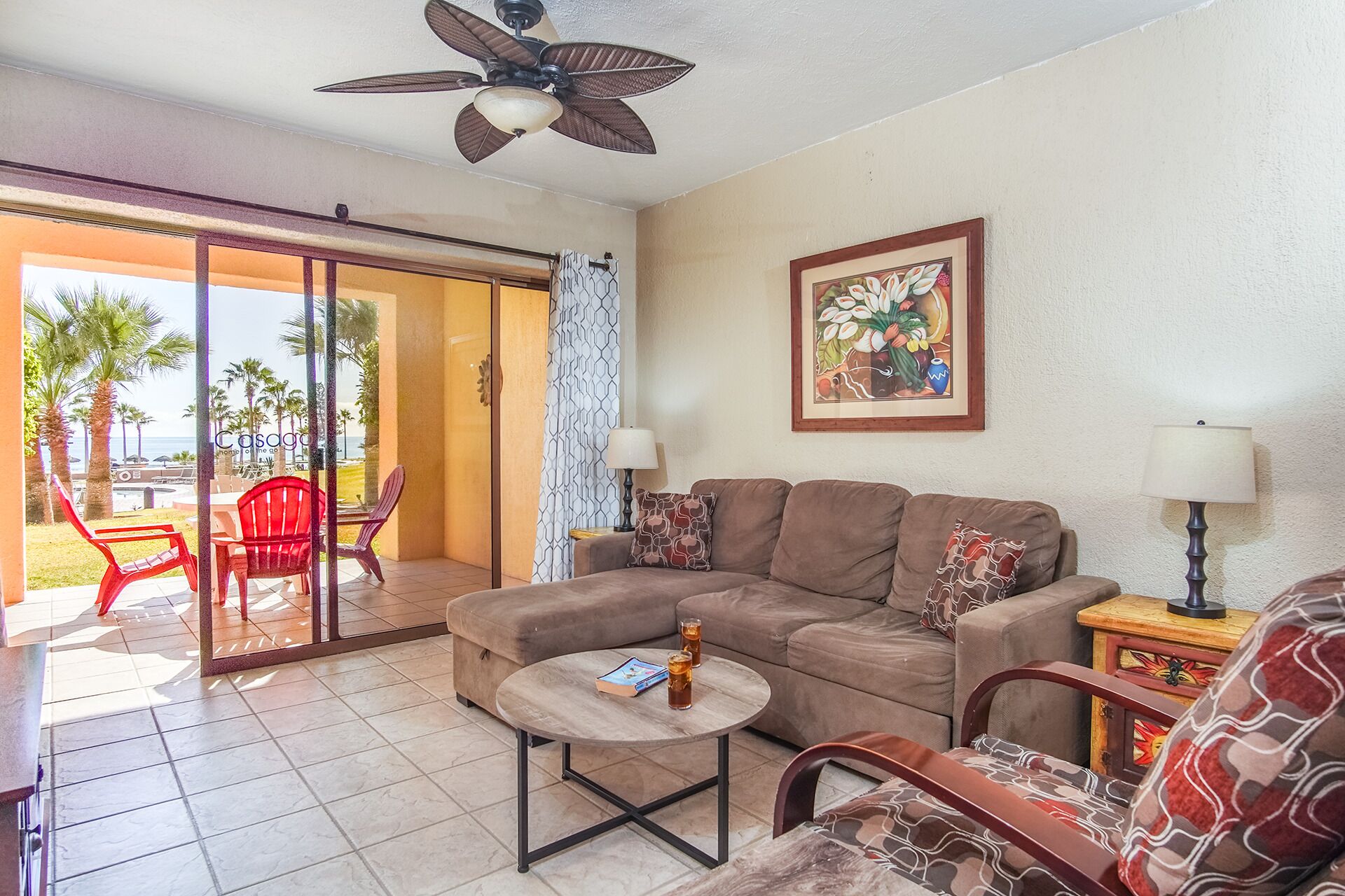 A living room with a sofa, coffee table, and chairs. There is a ceiling fan, a painting on the wall, and a view of the outdoors through a sliding door.