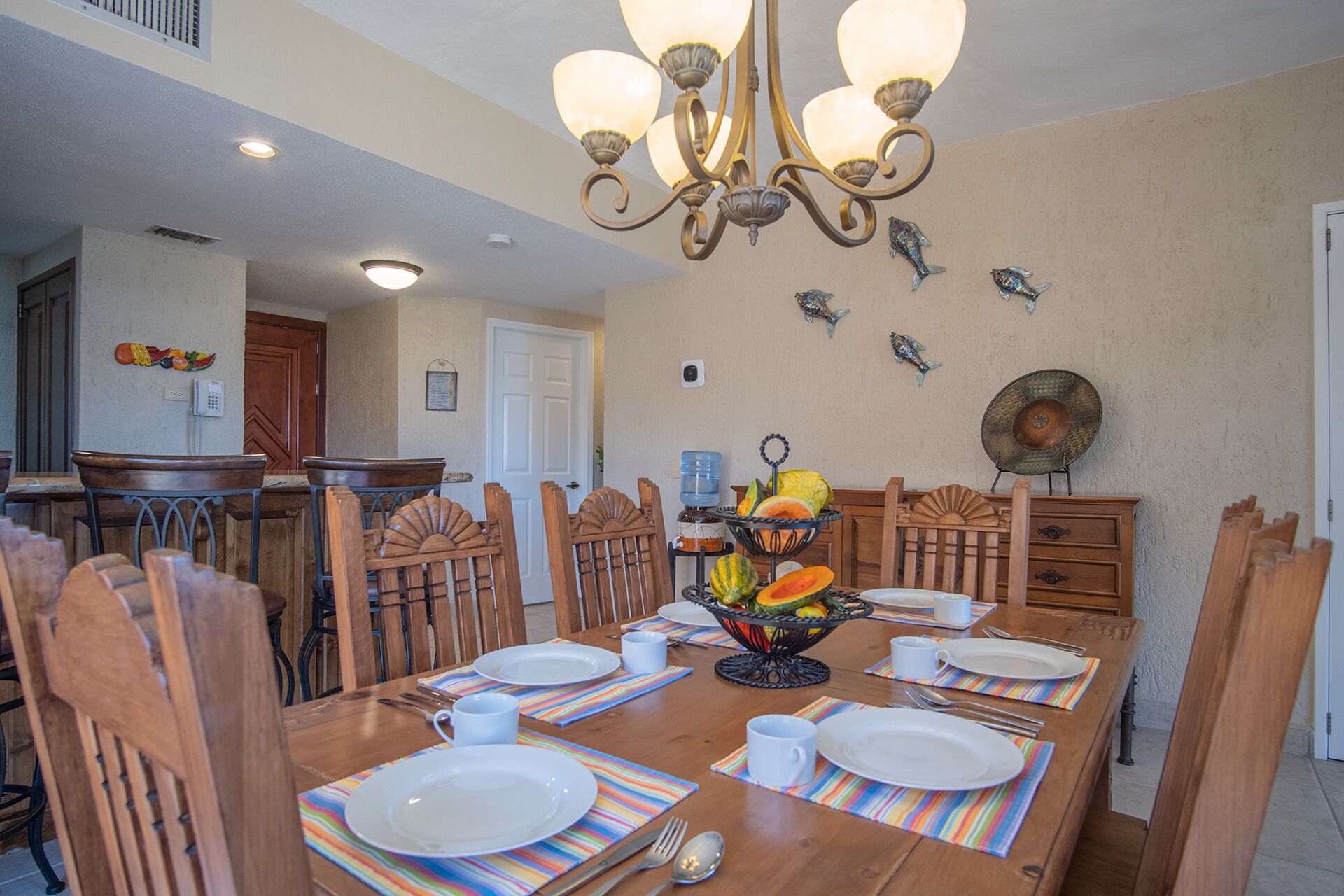 A dining area with a wooden table set for four, surrounded by chairs. A chandelier hangs above, and decorative fish are mounted on the wall.