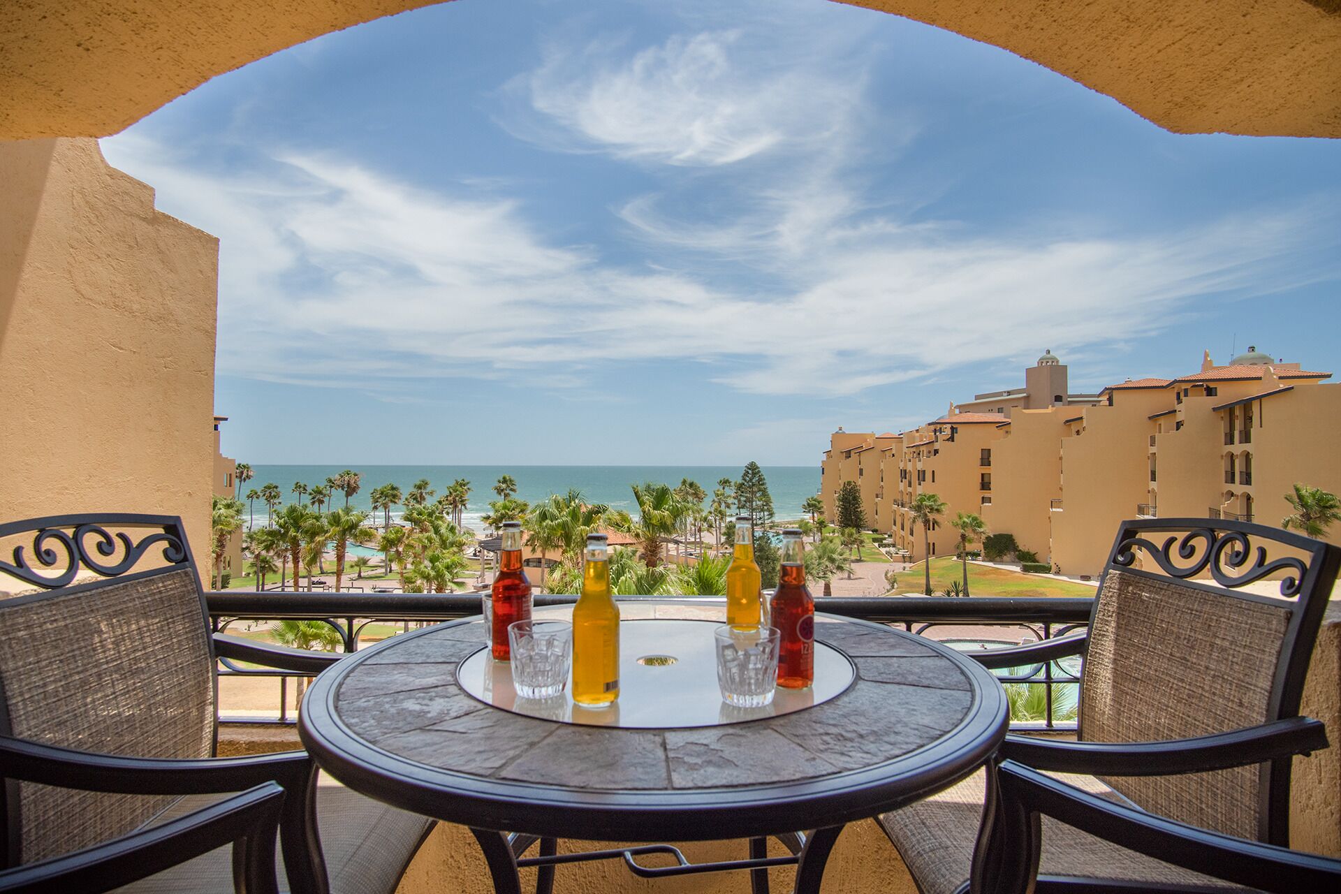 A balcony with a table, chairs, and bottles of beverages. A view of the ocean and palm trees is visible in the background.