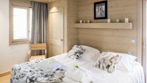 Une chambre confortable avec un plafond en bois, un lit avec une literie blanche et grise, une chaise, une fenêtre avec des rideaux et une étagère avec des bougies et un tableau.