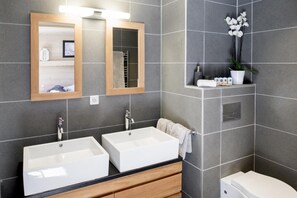 Ein modernes Badezimmer mit zwei Waschbecken, einem Spiegel, einem Handtuchhalter und einer Toilette.