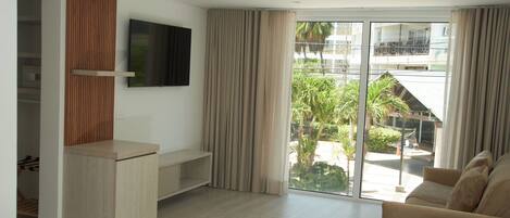 A modern living room with a flat-screen TV, a wooden entertainment unit, a beige sofa, and a sliding glass door leading to a balcony with a view of greenery and a building.
