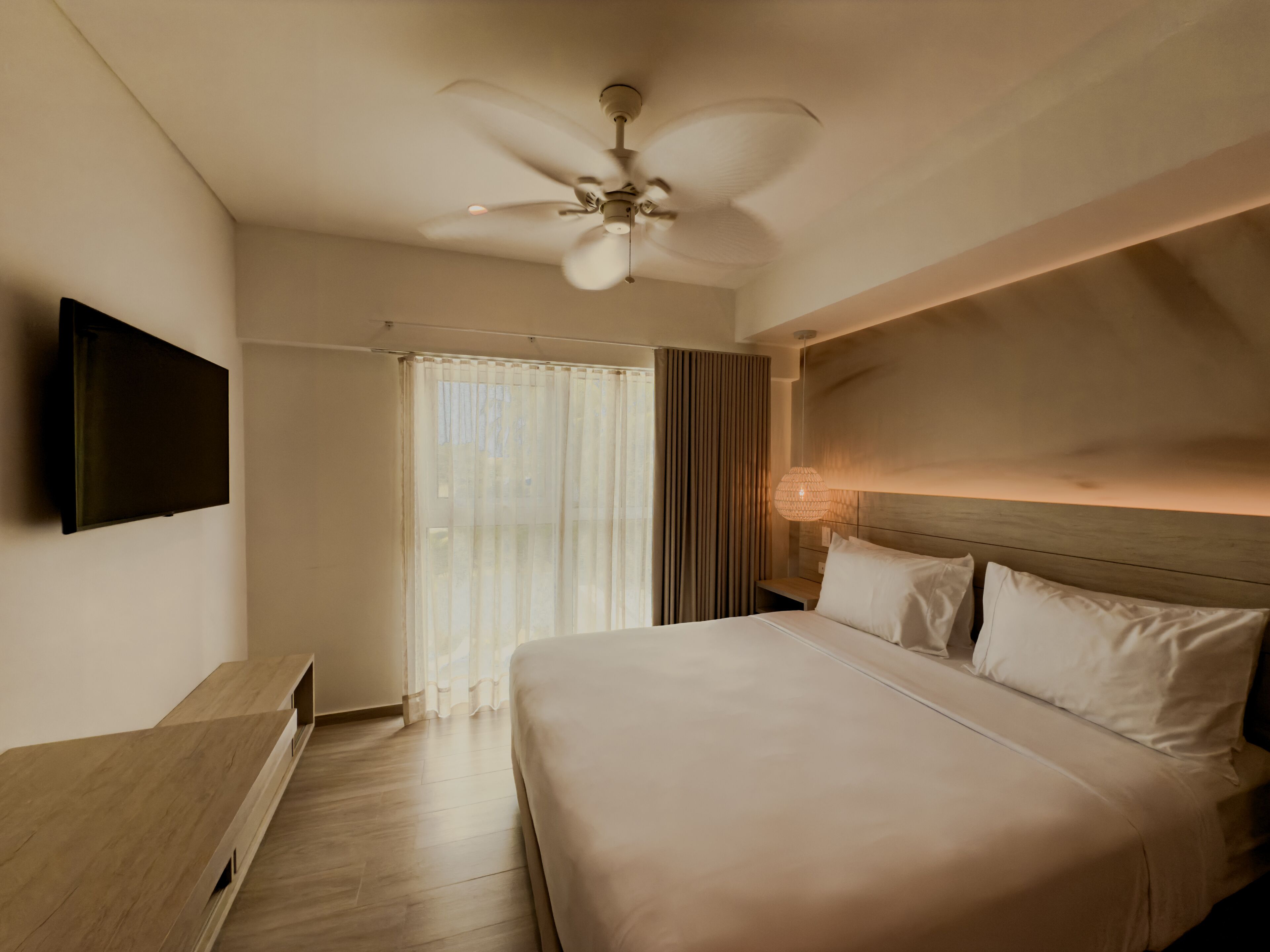 A hotel room with a bed, a ceiling fan, a TV mounted on the wall, and a window with curtains.