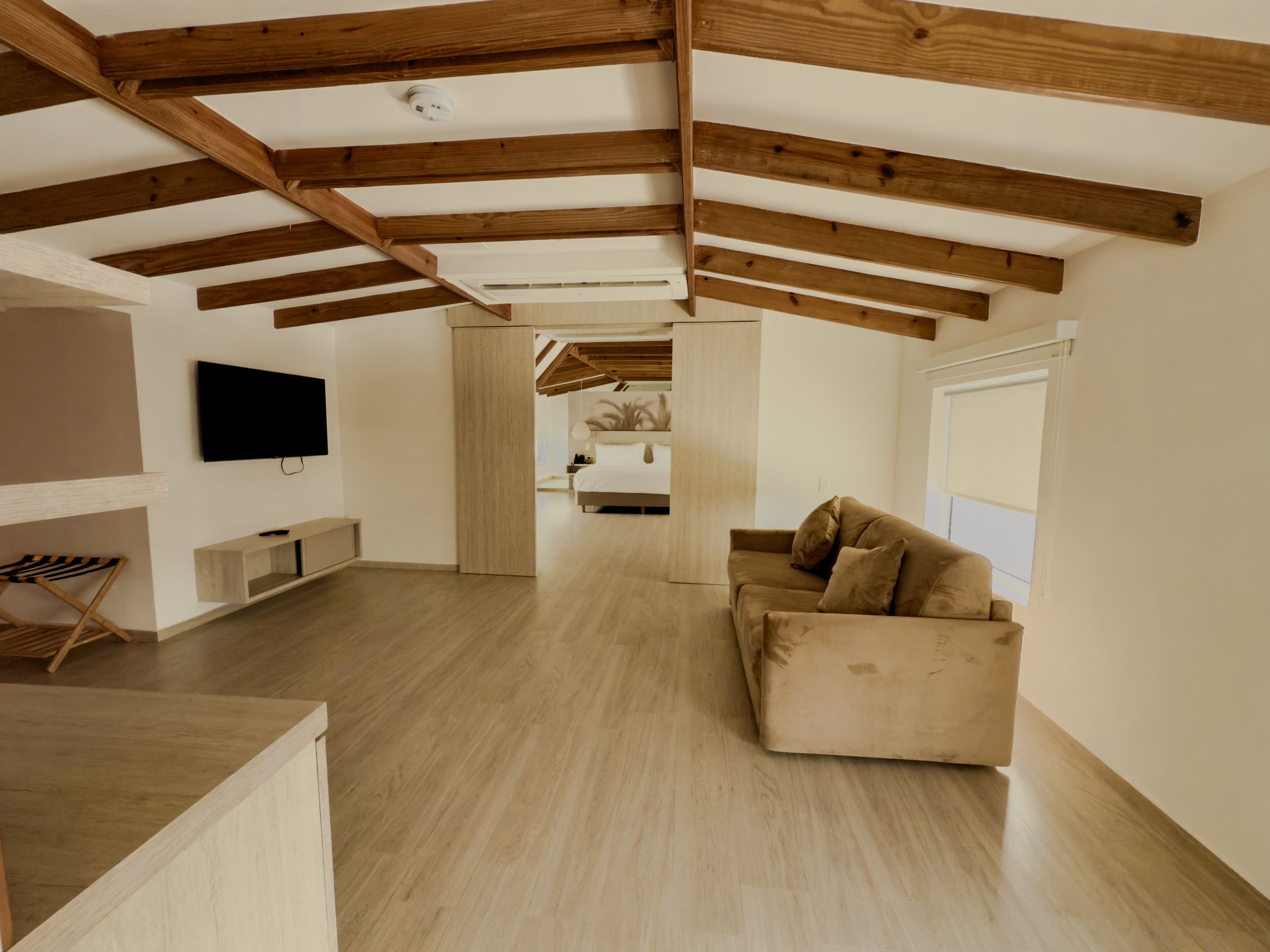 Un vaste espace de vie doté d’un plafond haut et de poutres apparentes, d’un téléviseur à écran plat fixé au mur, d’un canapé beige et d’un plancher en bois.