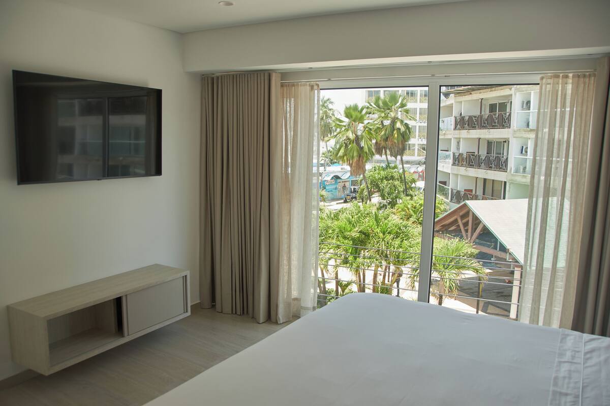 Una habitación de hotel con una cama grande, un televisor montado en la pared, una puerta corrediza de vidrio al exterior y un pequeño banco.