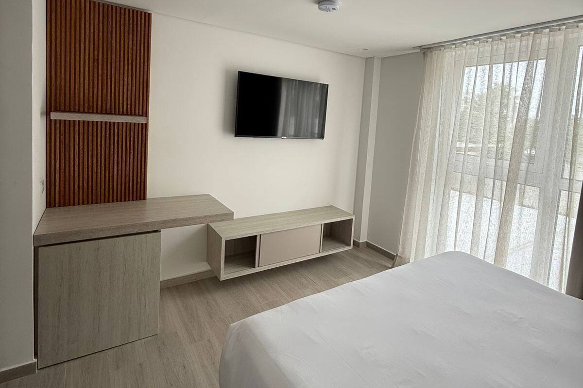 Una habitación de hotel moderna con una cama, un televisor montado en la pared, un panel de pared de madera y una ventana con cortinas traslúcidas.